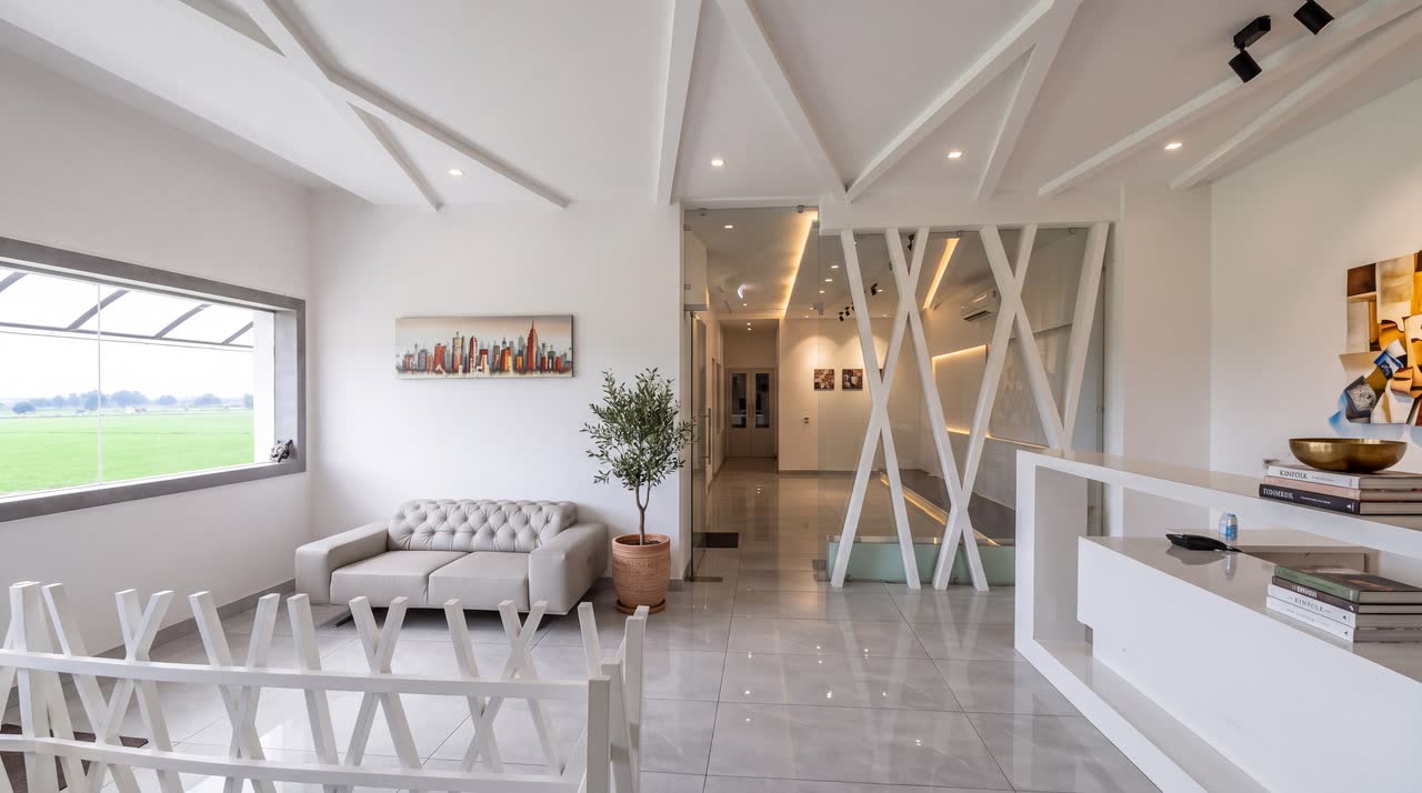 The entrance hall of the office: white walls and a white ceiling folded into angled beams, an open white screen beside the stair, a sofa under a window looking out to open field, and a reception counter on the right.